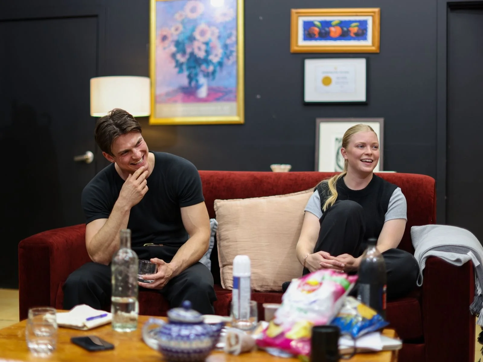Masterclass 2024 rehearsal: two students sit on a red couch in the studio, smiling, with snacks and drinks on a table in the foreground.