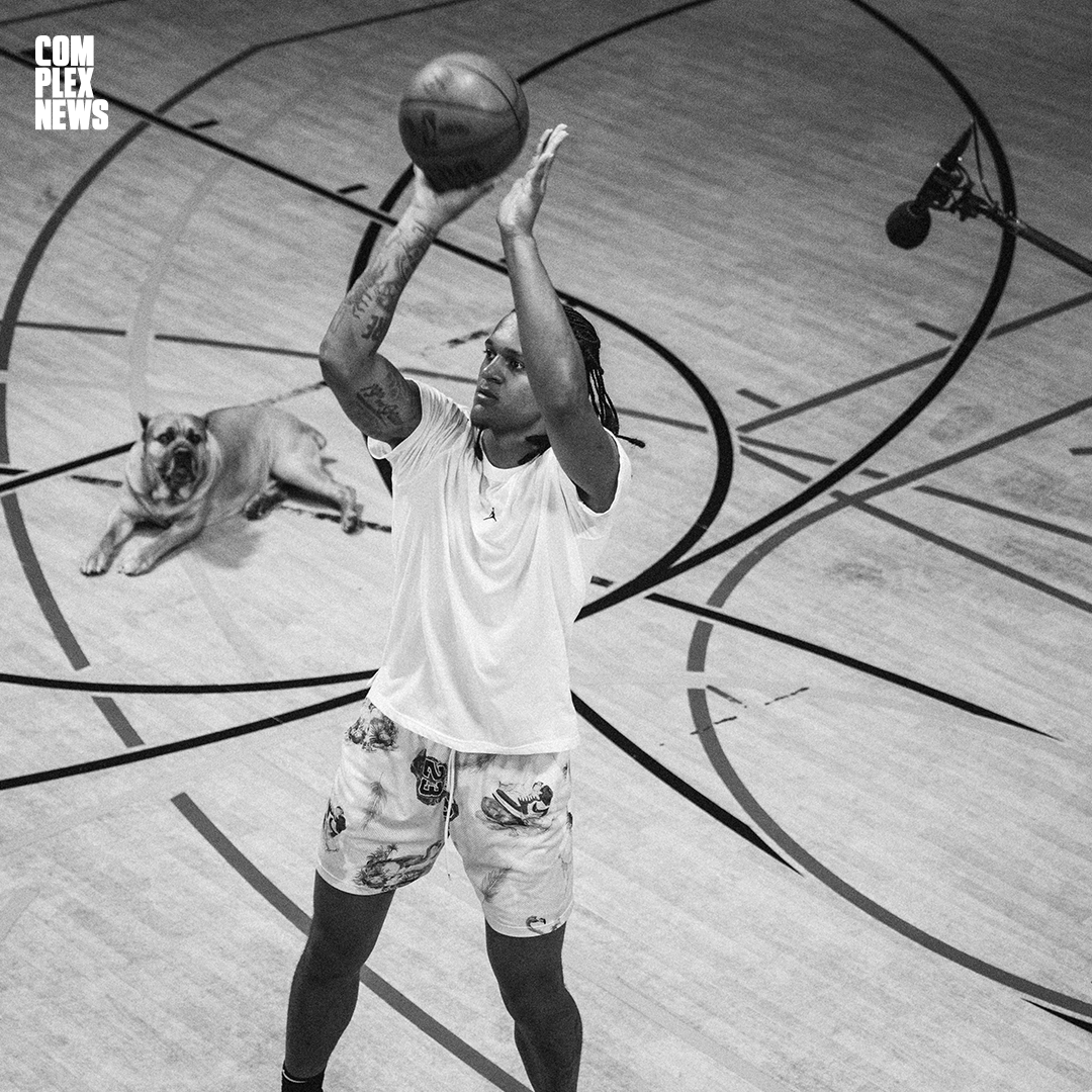 A basketball player with tattoos on his arm is shooting a shot on an indoor basketball court, with a bulldog laying on the court behind him.