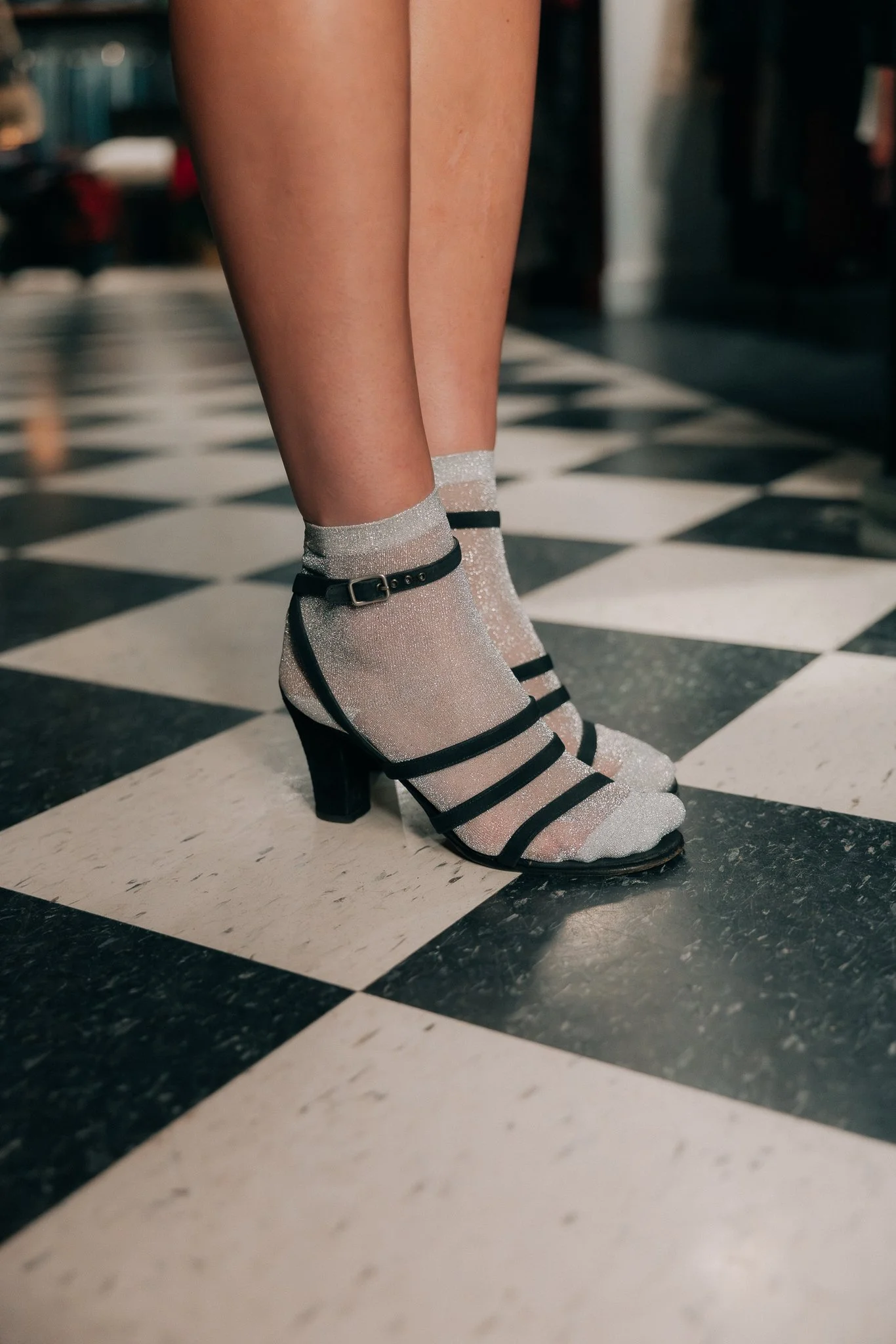 Close-up of a person's legs wearing glittery silver high-heeled shoes with black straps and ankle buckle, standing on a black-and-white checkered floor.