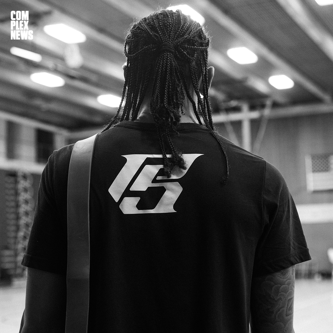 Back of a person with braided hair wearing a black T-shirt with a large logo on the back, standing indoors with a high ceiling and bright lights.