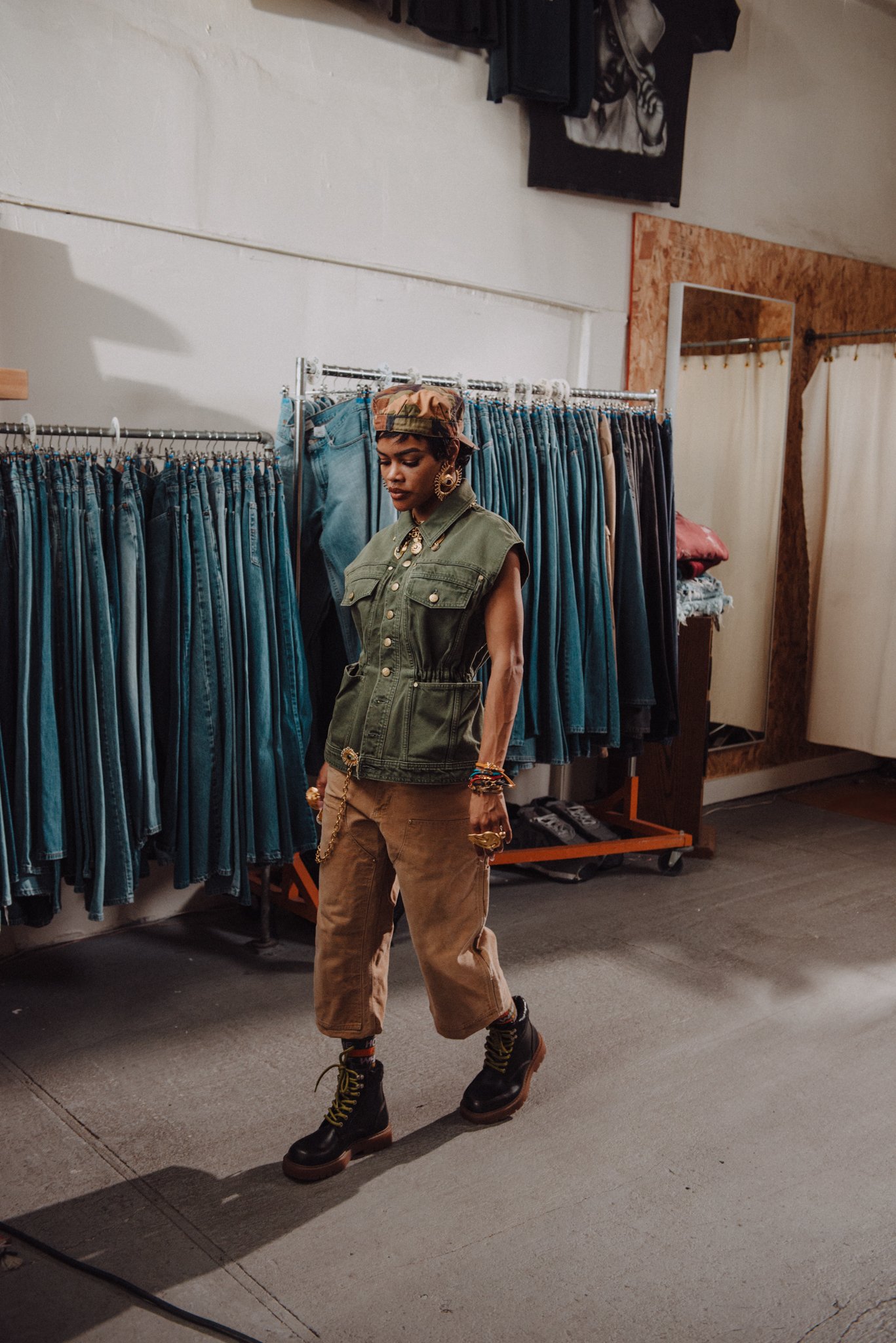 A person with dark skin and short hair, wearing a camo cap, green sleeveless utility vest, brown cargo pants, and black combat boots, walking in front of a clothing rack filled with blue jeans in a store.