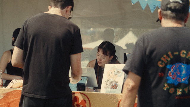 People at a vendor booth with a girl working behind the counter, and two men standing in front. The booth has a sign with colorful artwork.