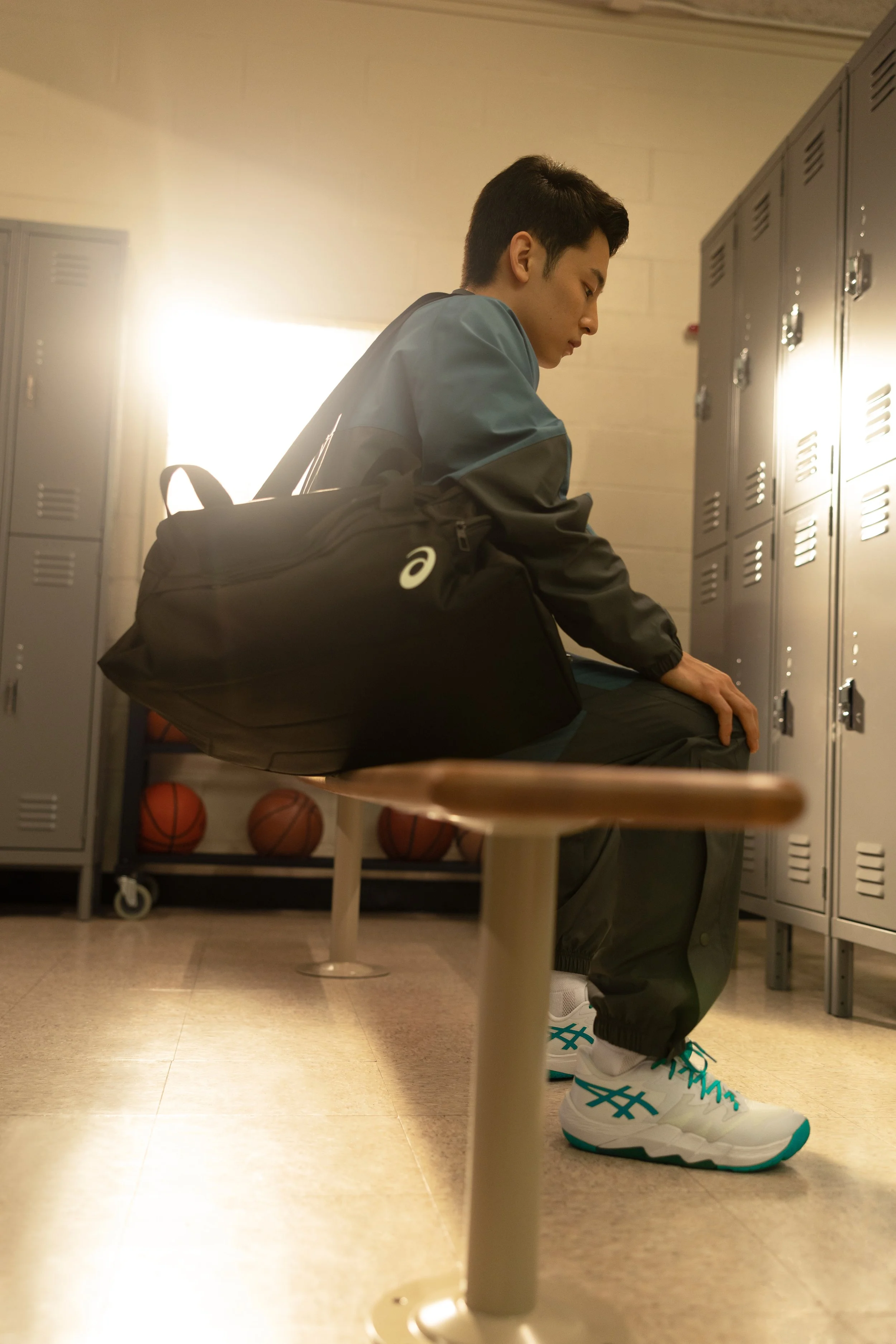 A young man sitting on a school bench in a locker room, with lockers and basketballs in the background, wearing athletic shoes, carrying a backpack, and looking down.