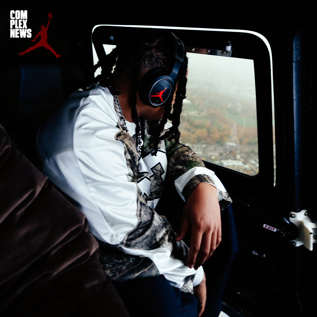 Person wearing a white and camouflage jacket, headphones with a red Jumpman logo, sitting in a helicopter with a view of the landscape outside.