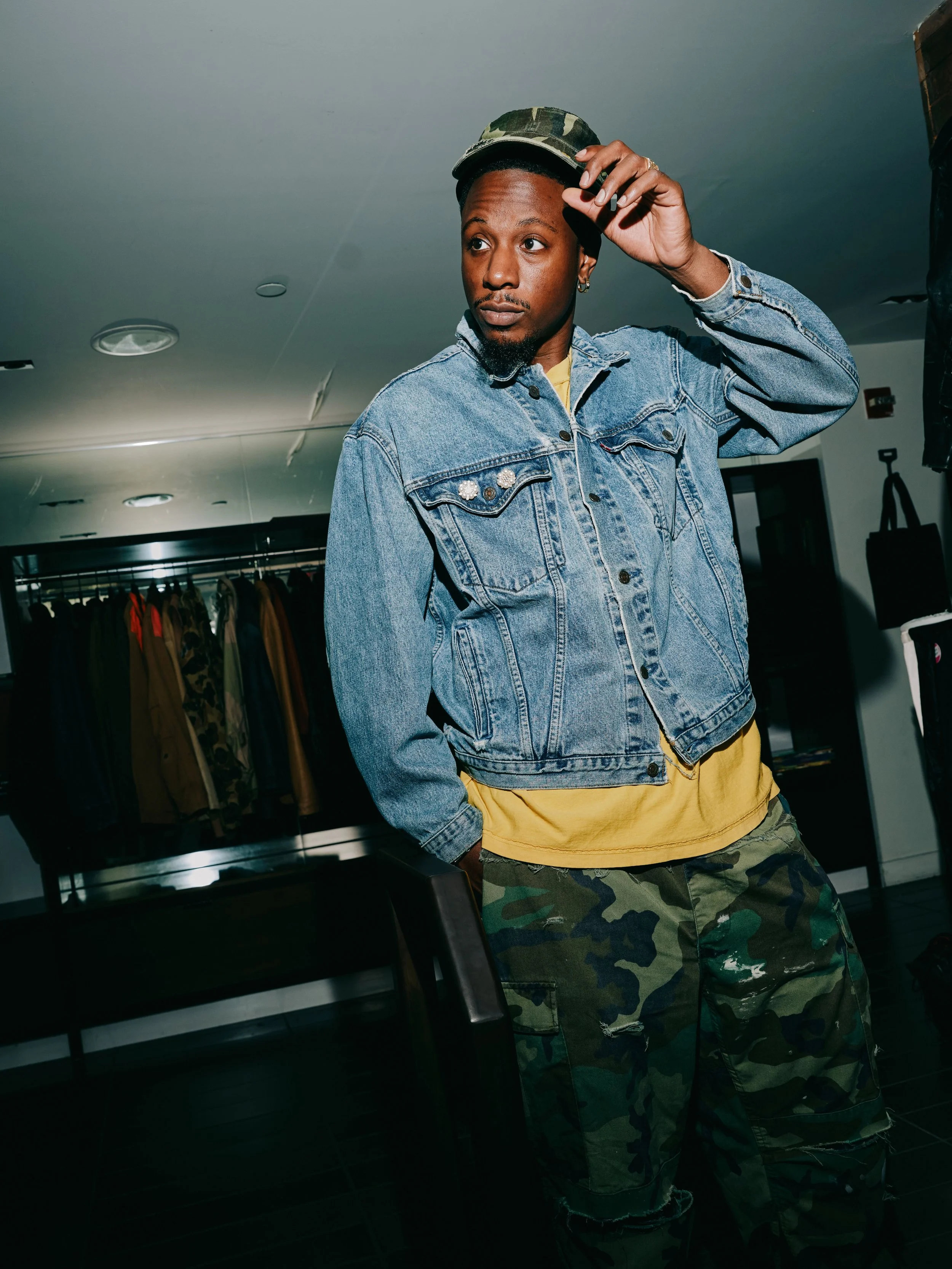 A man wearing a camouflage hat, denim jacket with pearl embellishments, yellow shirt, and camouflage pants, standing in a clothing store. He is adjusting his hat with one hand, standing in front of clothing racks, with a contemplative expression.