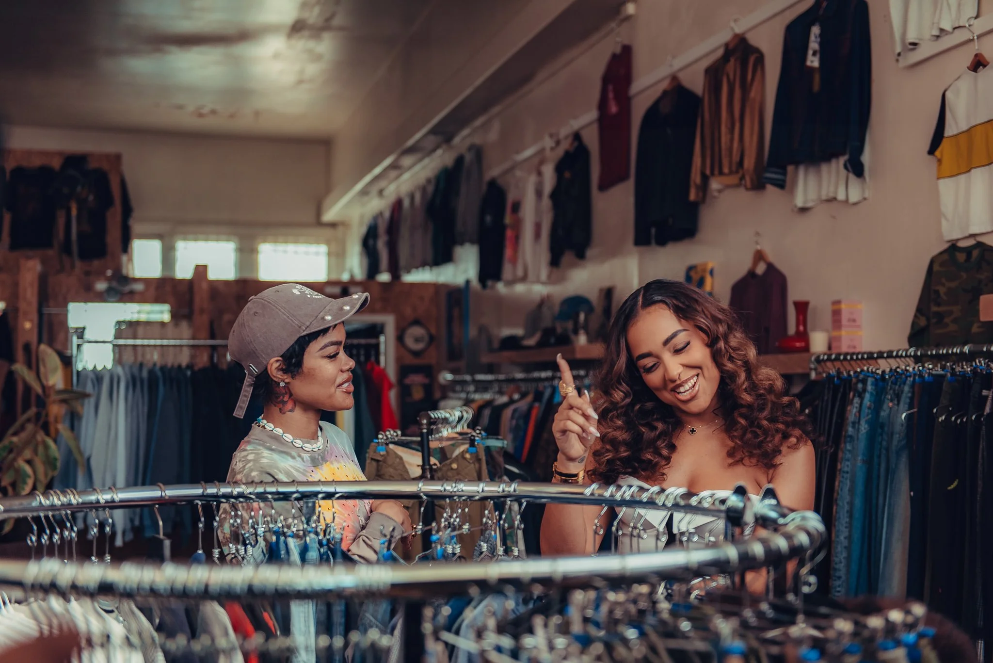 Two women shopping in a clothing store, looking at racks of clothes, with one woman smiling and the other wearing a cap.
