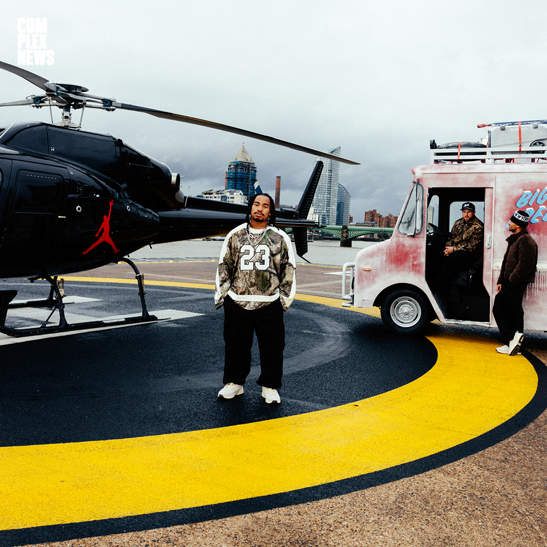 A man standing on a helipad near a black helicopter with a red Jumpman logo, a pink food truck, and two men in military and casual attire in front of a city skyline with cloudy skies.