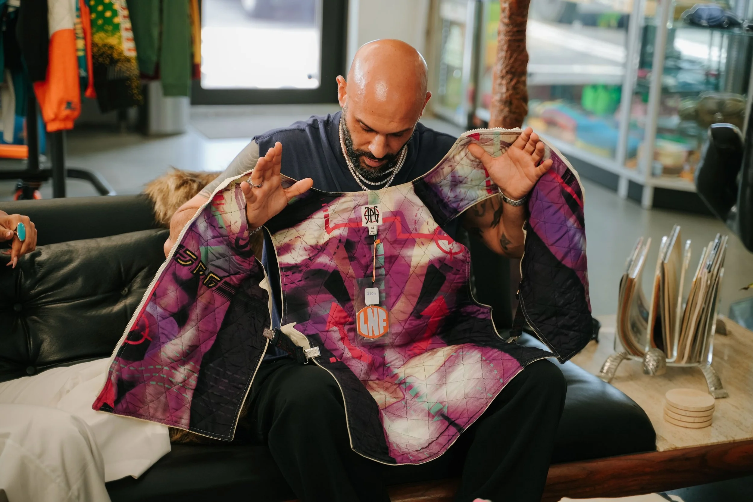 A bald man with a beard is sitting on a black leather couch inside a store, holding a colorful patterned jacket and looking down at it. The store has clothing items hanging on racks, and a door with glass panels is visible behind him.