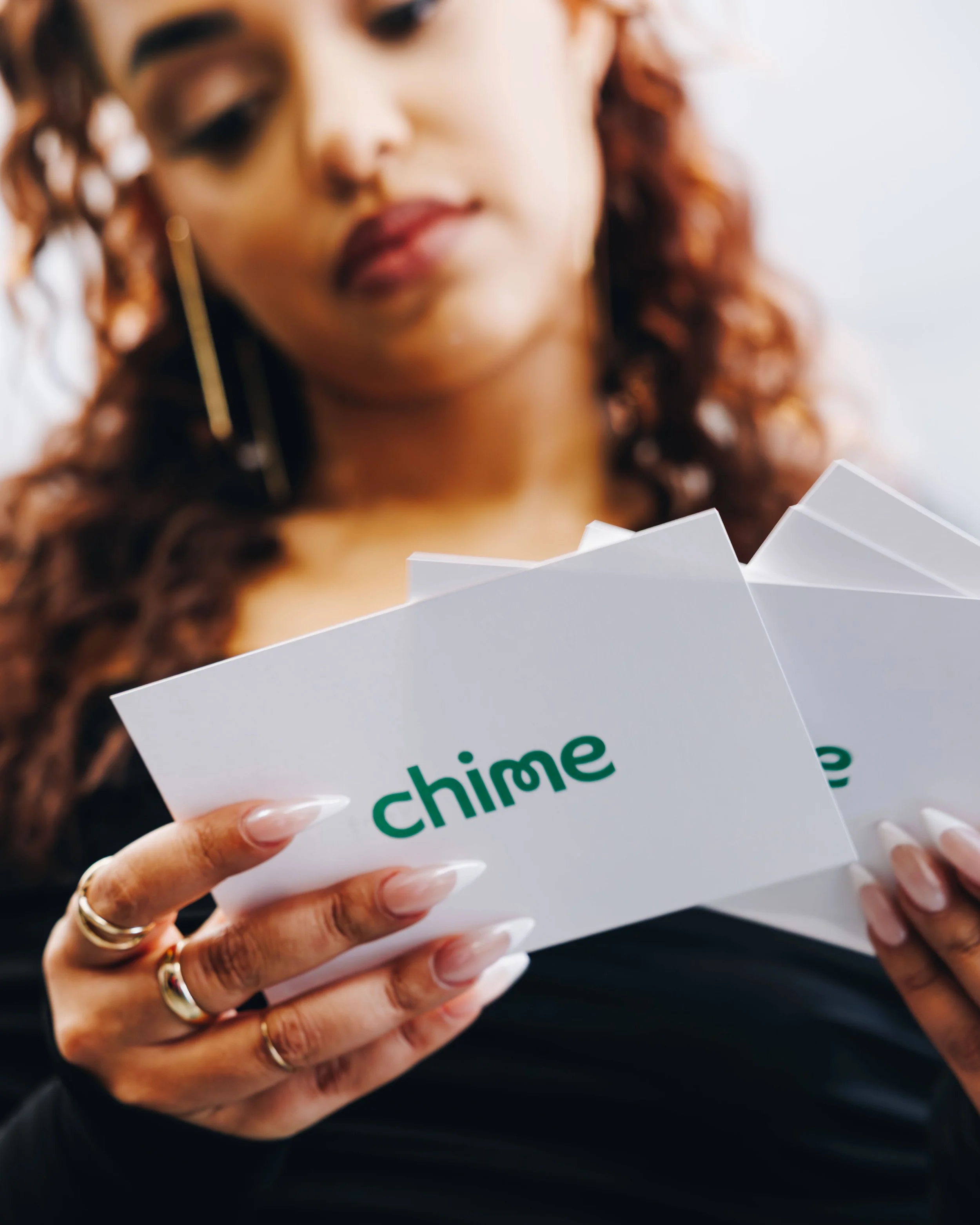 A woman with long, wavy, reddish hair and a gold earring holds a white box with green text that reads 'Chime.'