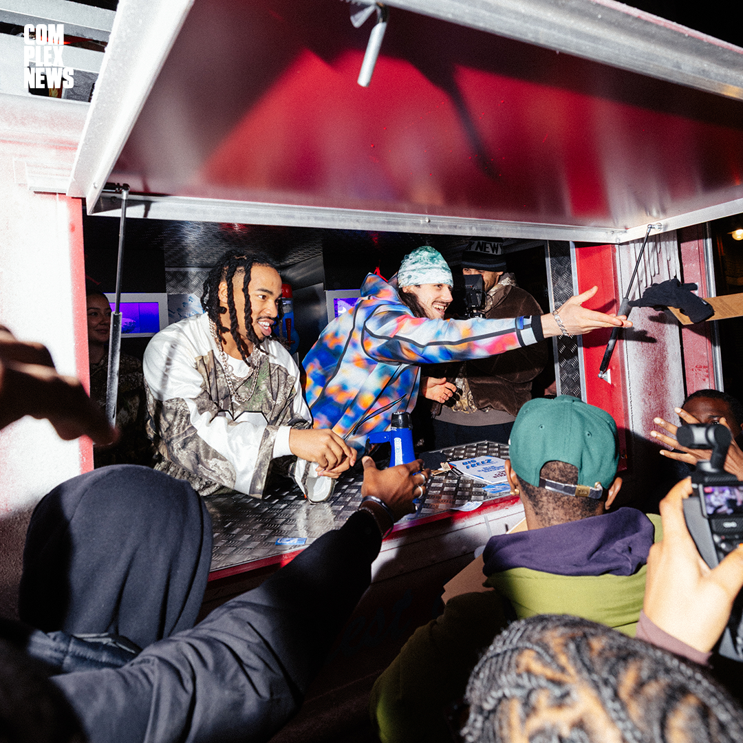 Two men at a food truck, one with dreadlocks and the other wearing a tie-dye jacket and beanie, interact with a crowd at night.
