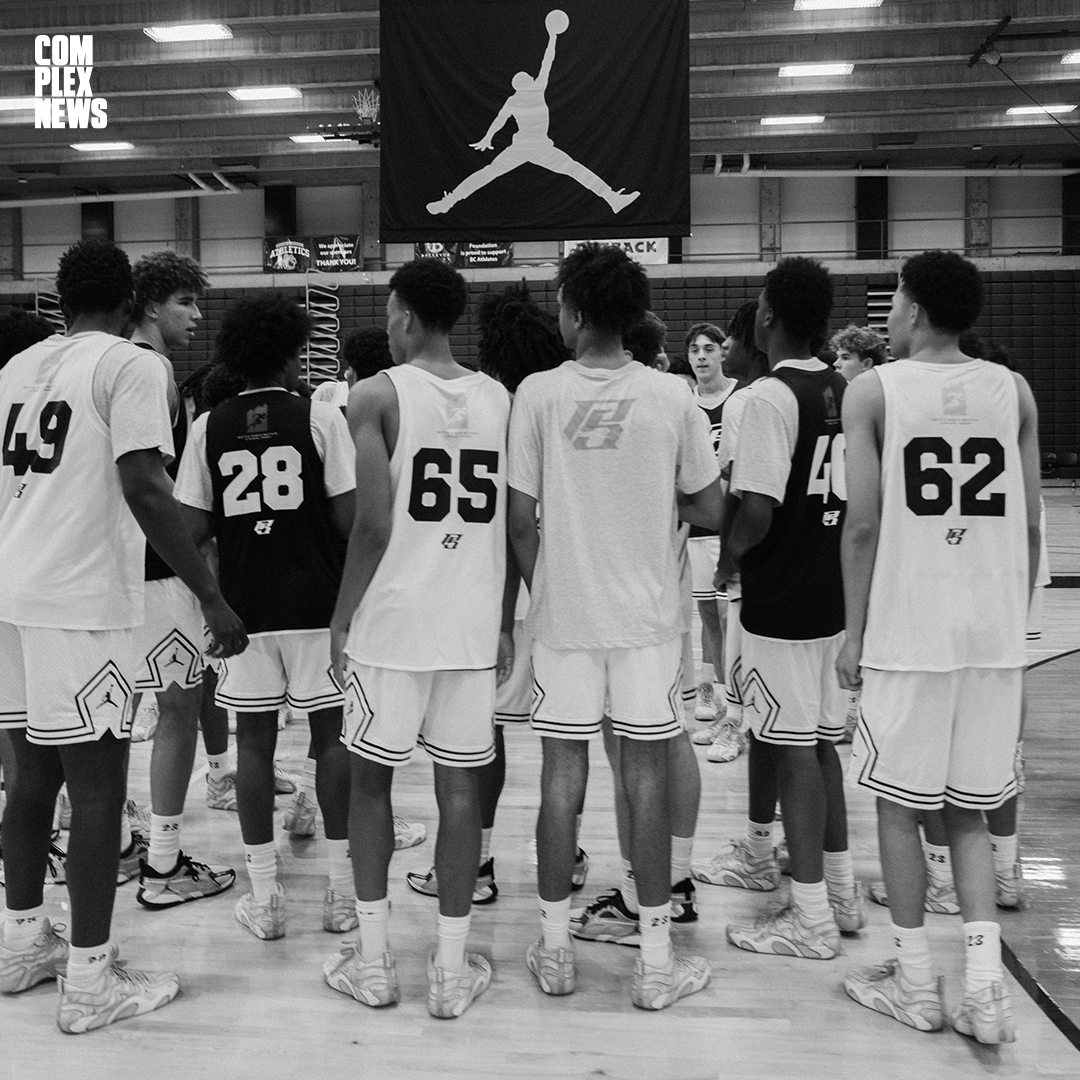 A group of young basketball players in sports uniforms gathered in a gymnasium, listening to a coach or team leader. The gym has a large Jordan brand logo banner hanging from the ceiling.
