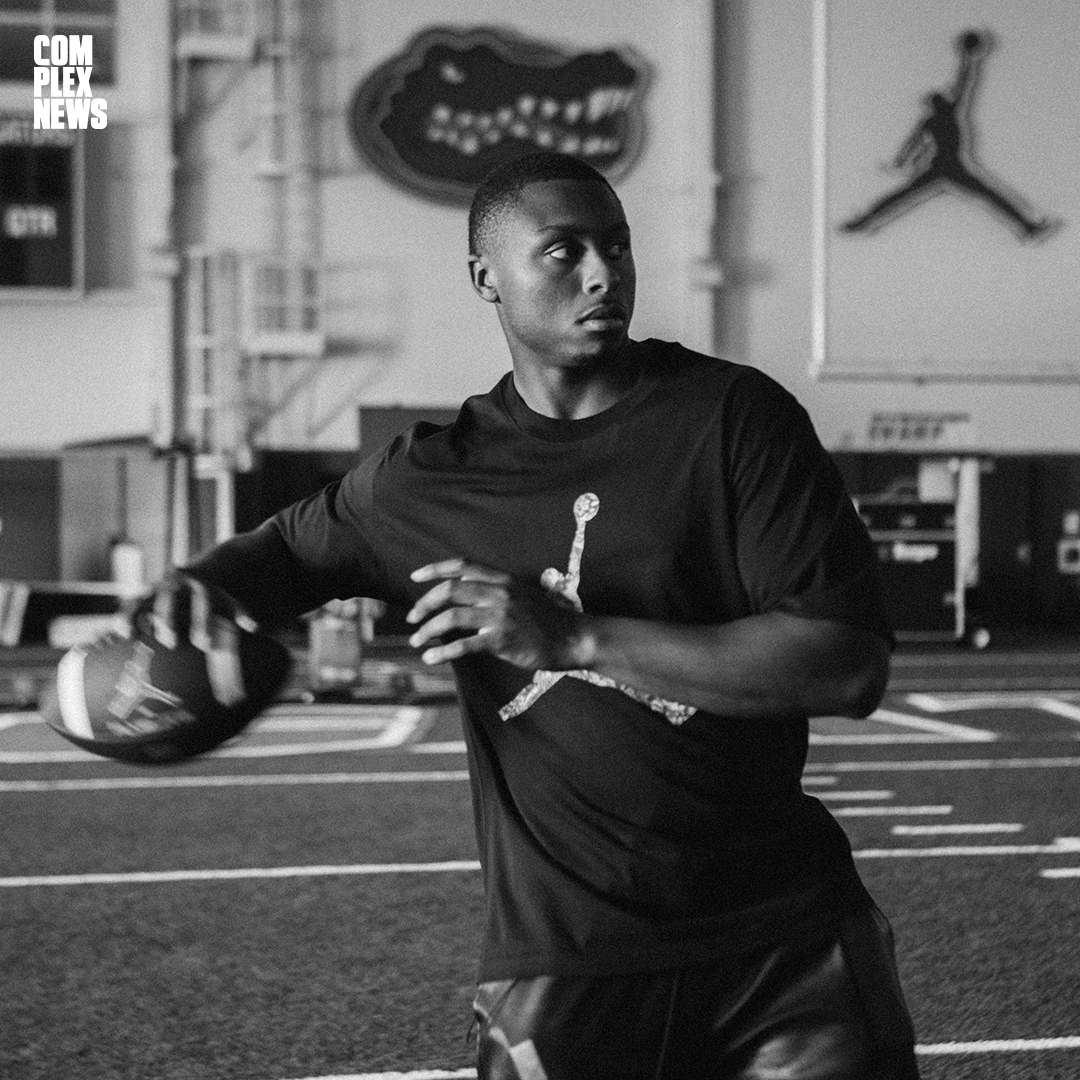A young man in a black t-shirt and shorts throwing a football in an indoor sports facility with sports logos on the wall.