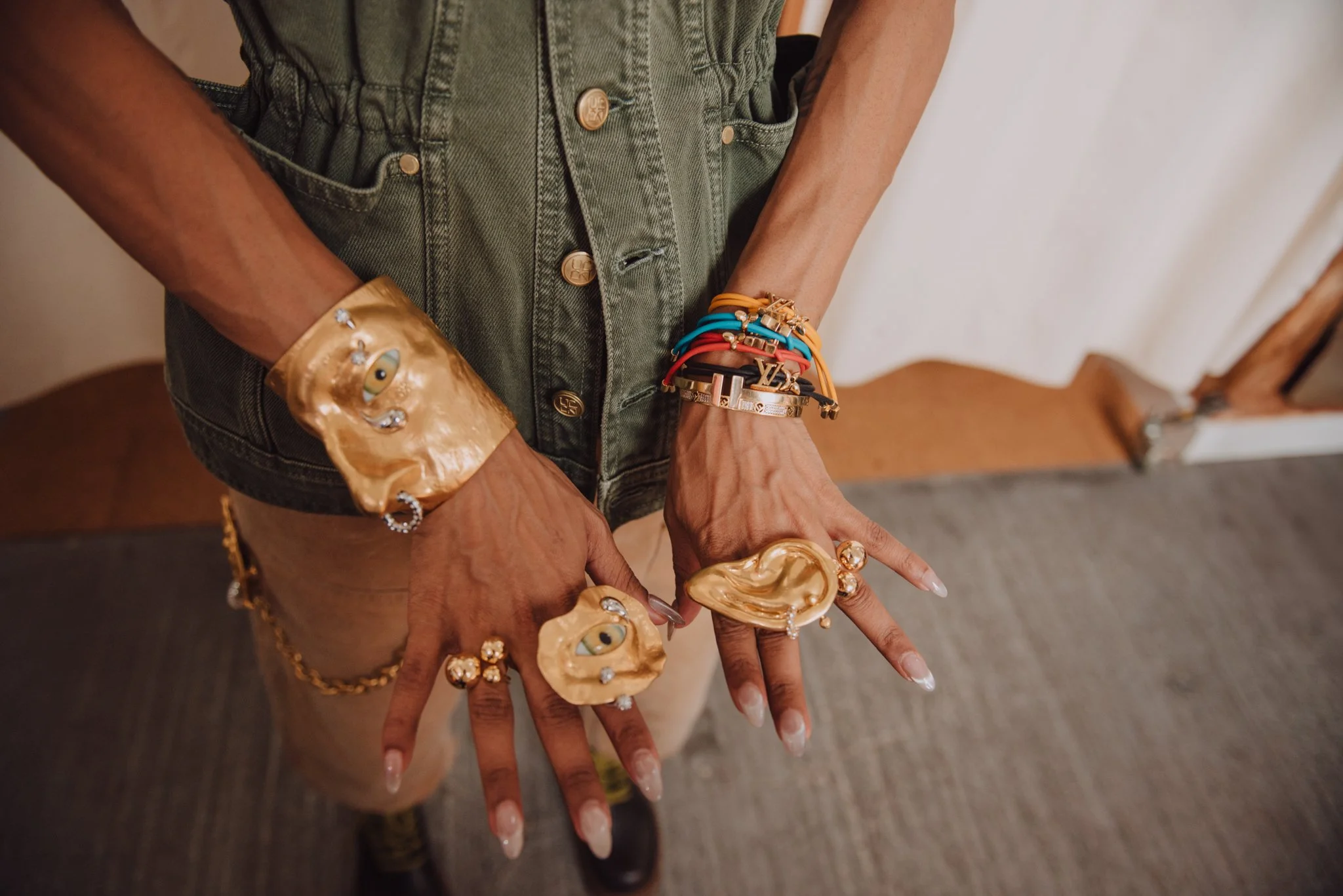A person wearing a green denim vest with multiple bracelets and rings on their hands, with long manicured nails, showing off their jewelry and accessories.