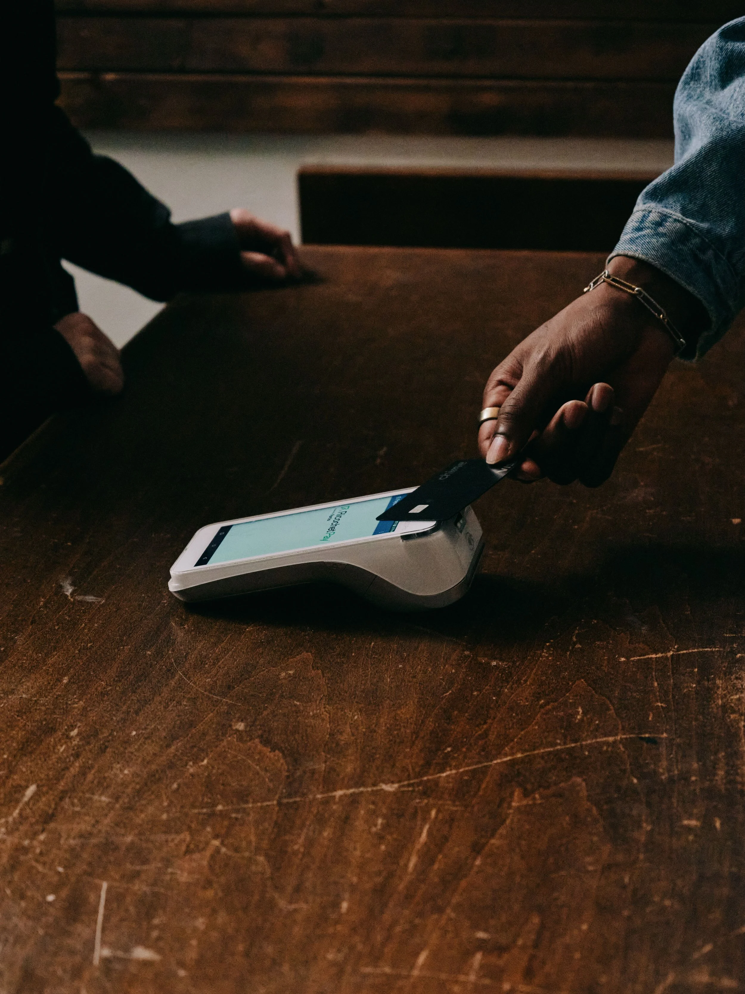 People exchanging a contactless payment using a smartphone and card reader on a dark wooden table.
