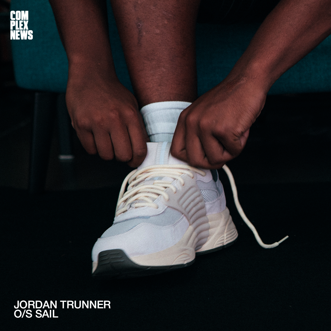 Person tying the laces of a white Nike Jordan Trunner O/S Sail sneaker, wearing white socks, against a dark background.