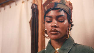 Close-up of Teyana Taylor wearing a camouflage cap, gold earrings, and a green jacket, standing in front of beige curtains.