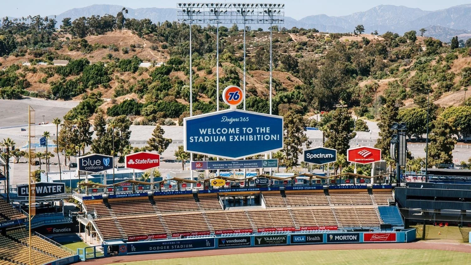 Empty baseball stadium with signs that say 'Welcome to the Stadium Exhibition,' sponsor logos, and scenic hills in the background.