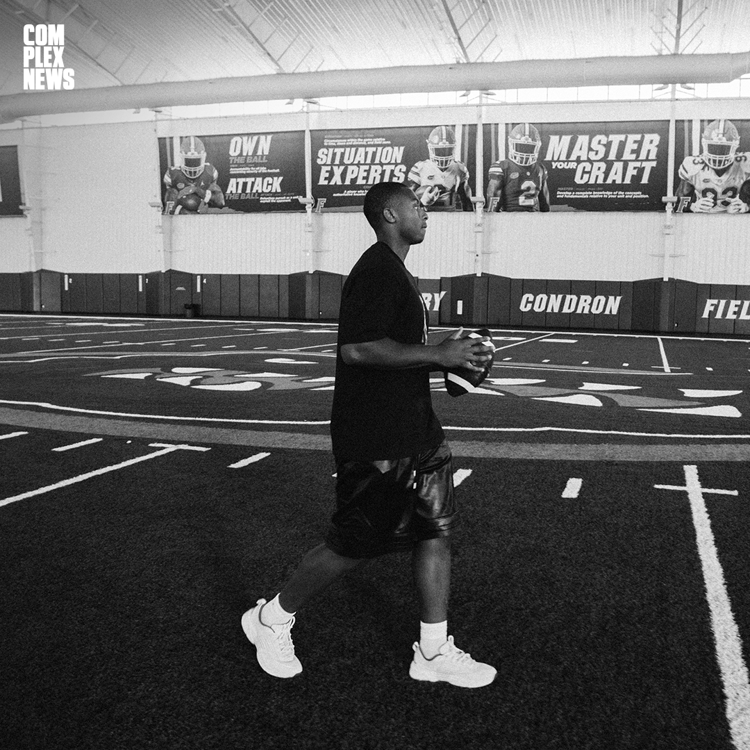 A young man is walking inside an indoor football field, holding a football. The field has painted yard lines and a track surrounds it. There are large posters on the wall with football players and motivational phrases.
