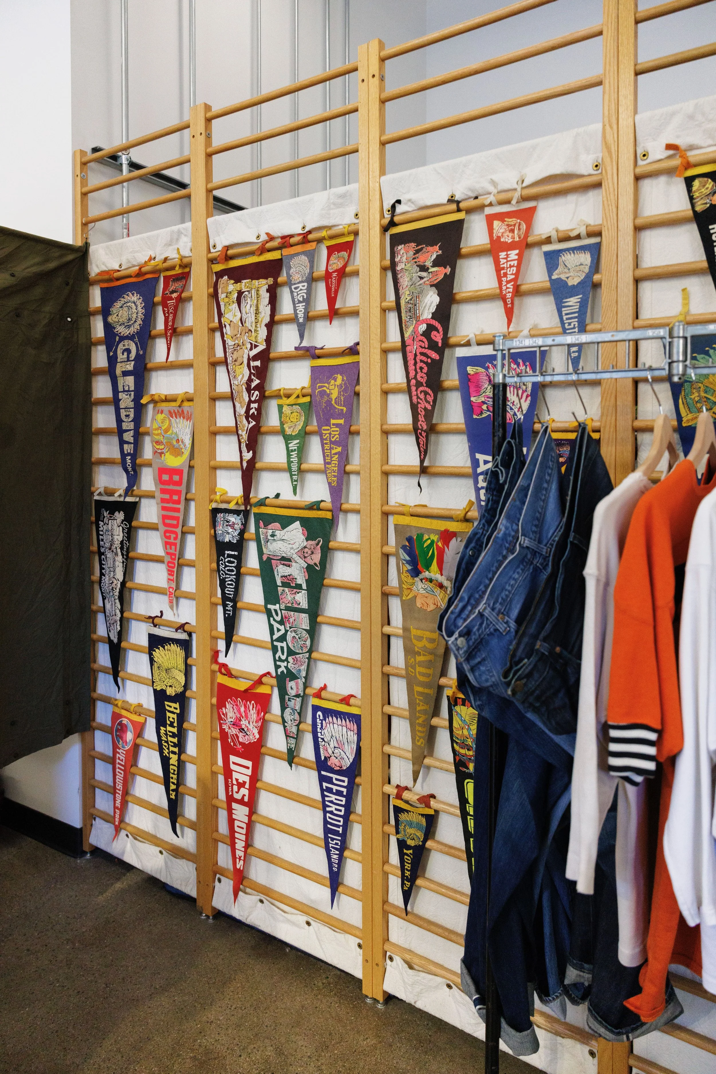 A display of colorful pennant banners hanging vertically on a wooden wall rack inside a room, with some clothing items like jeans and shirts hanging nearby.