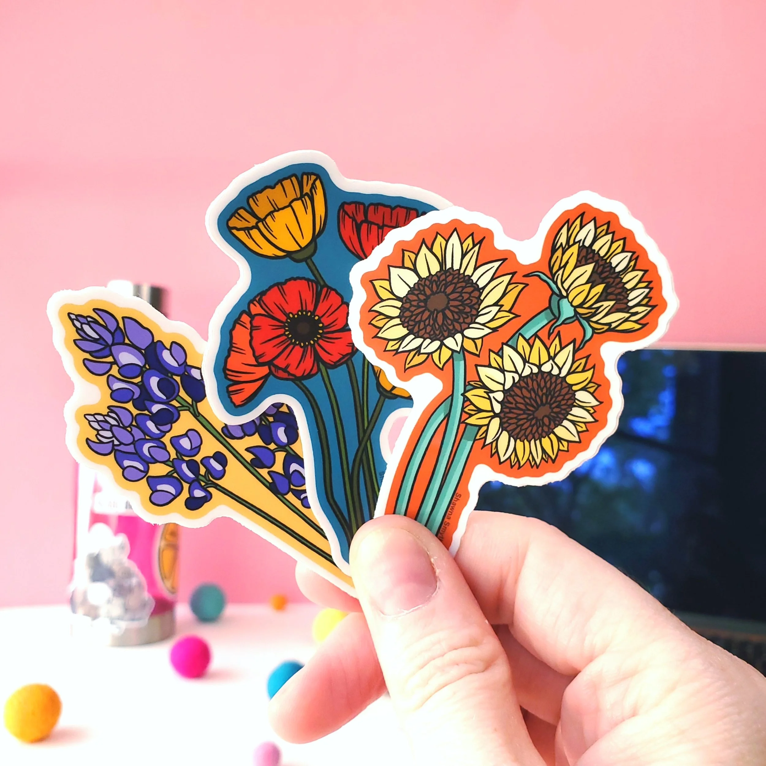 Close up of a woman's hand holding a trio of flower stickers- one sunflower, one poppy, one bluebonnet- in front of a decorative plant and a water bottle.