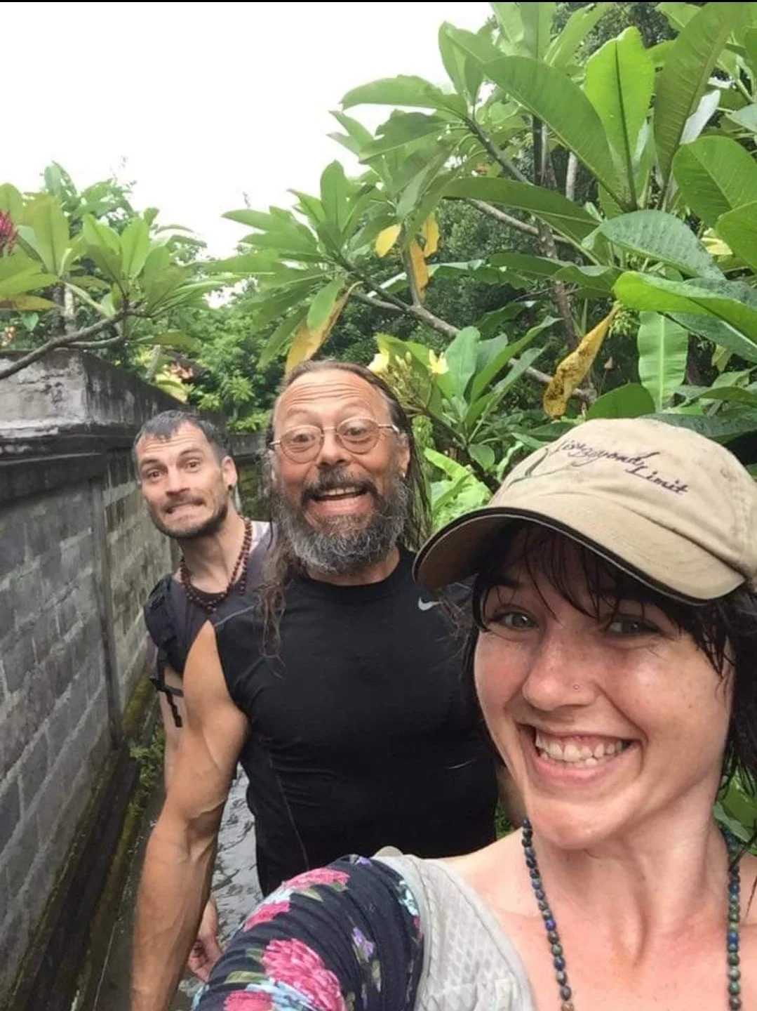 One Woman with a Live Beyond Limit hat and Two bearded men smiling in a line on a path between tropical plans and a cinderblock fence line in Bali, Indonesia.