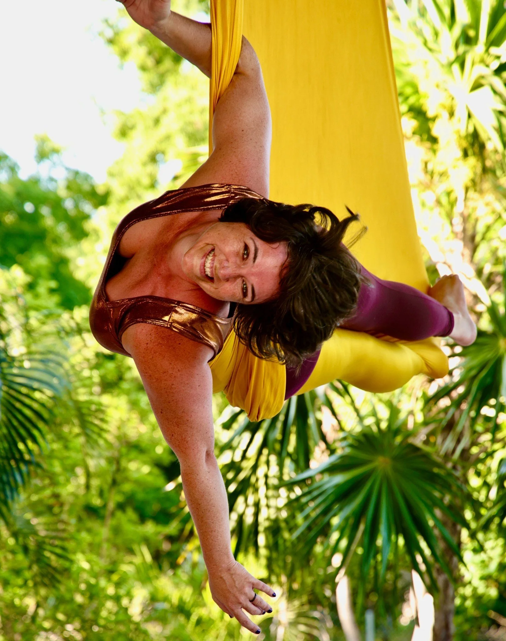 Yoga Teacher in yellow aerial hammock holding with one arm and with her legs wrapped while smiling in front of tropical foliage