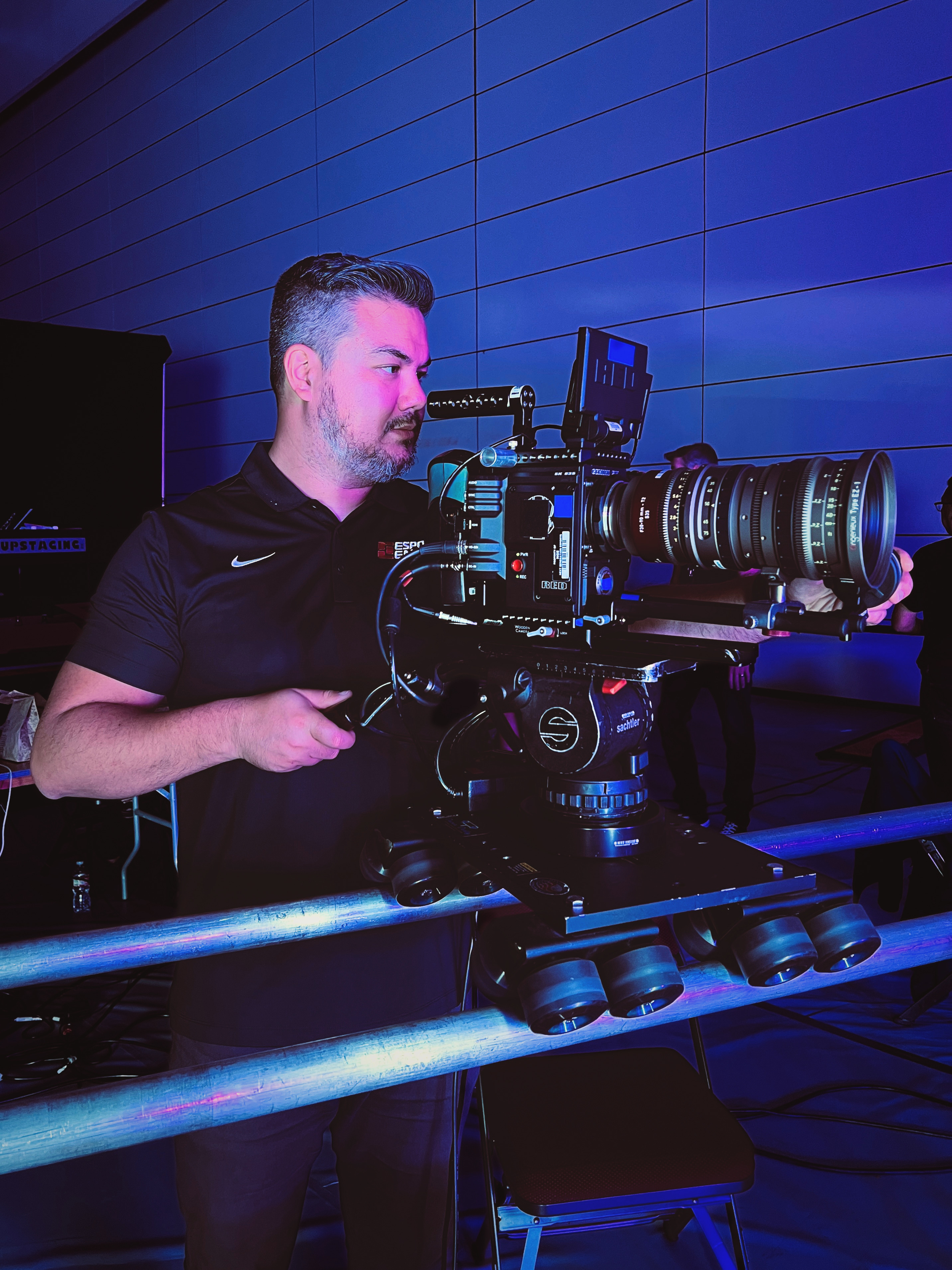 A man operating a large professional film camera with a long lens at an indoor event, with a blue-lit backdrop and a few people in the background.
