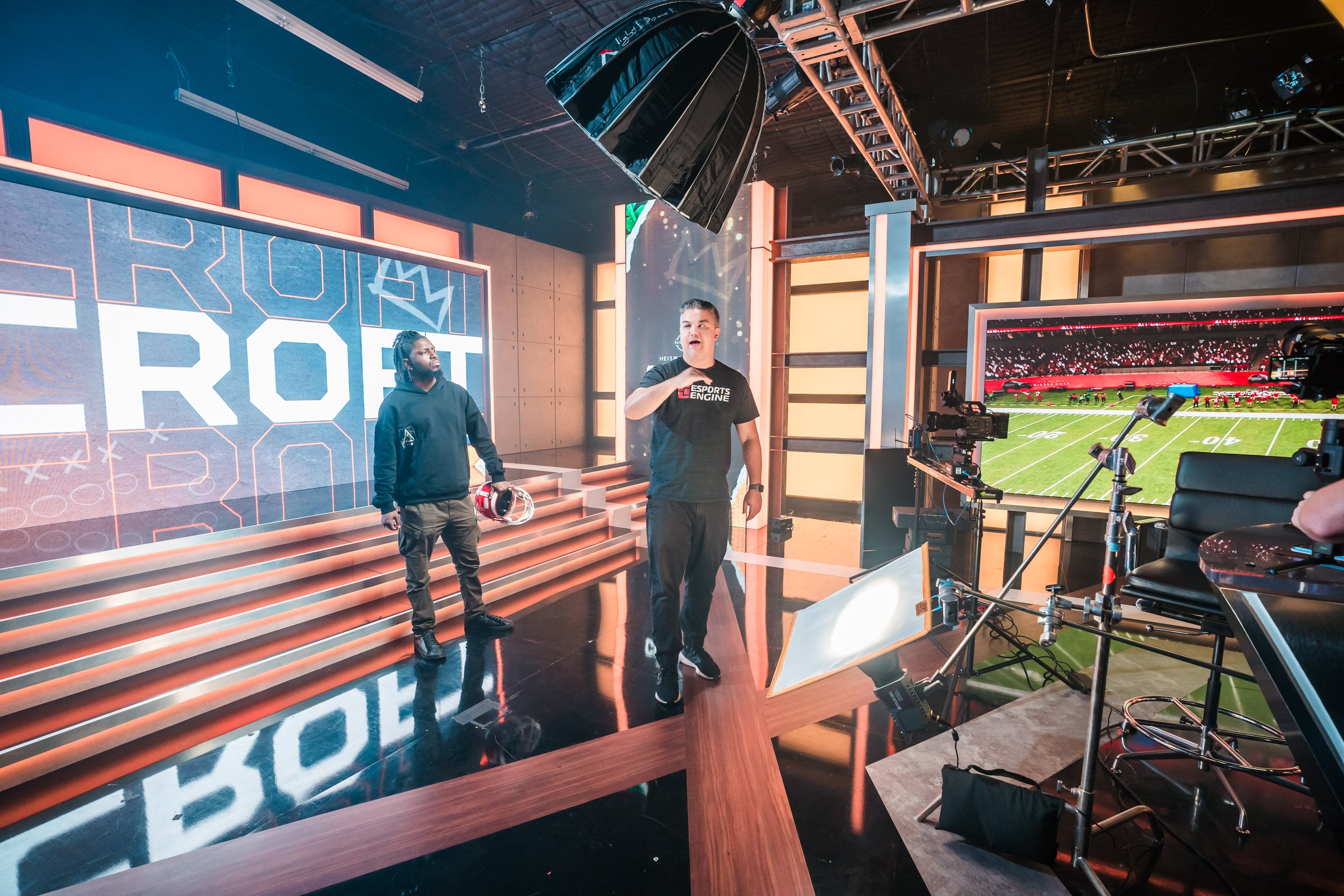 TV studio with two men preparing for an upcoming esports broadcast, one speaking, with camera equipment and a football field displayed on a large screen in the background.