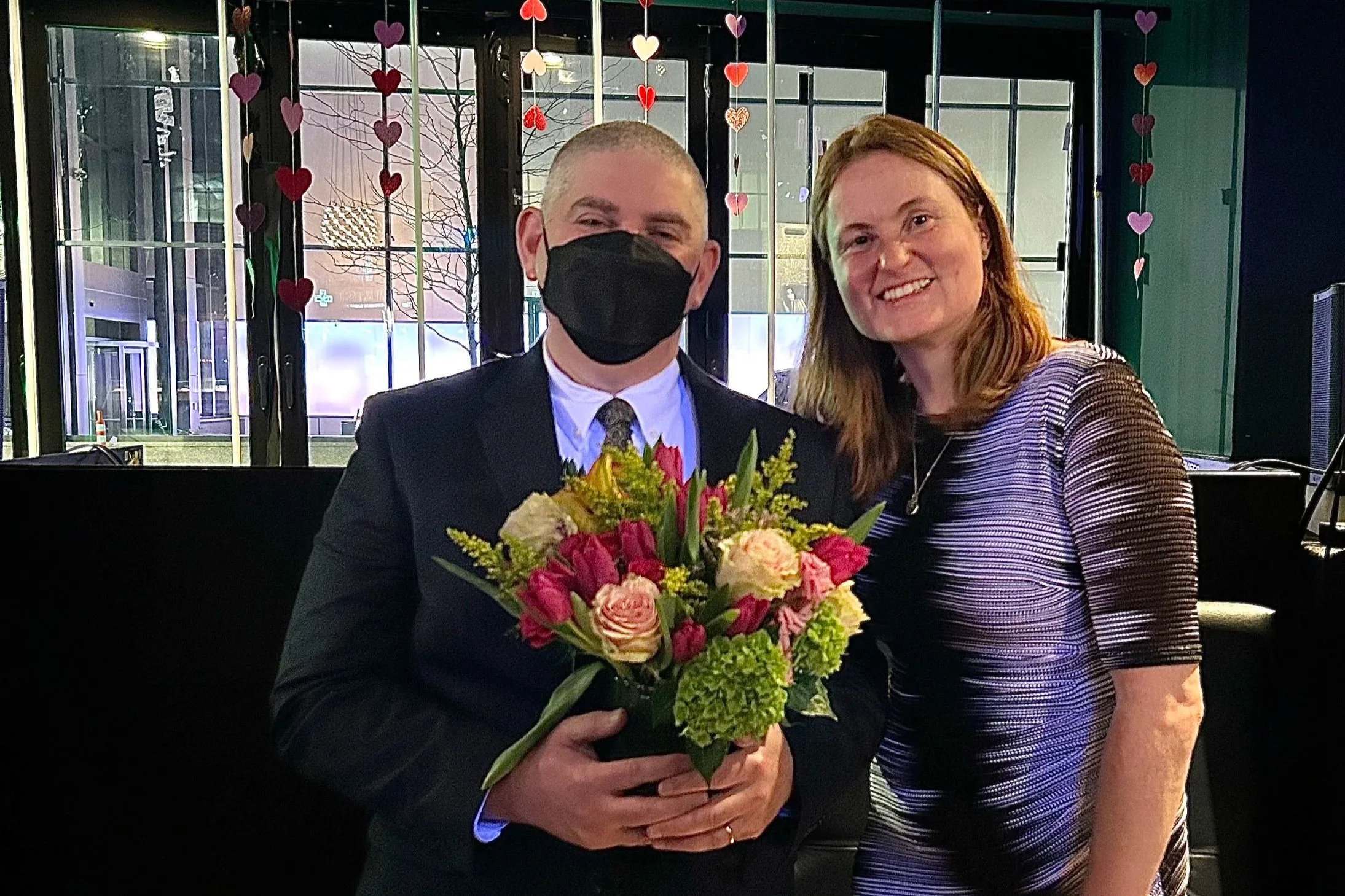 Attorney Nathanson stands next to Attorney Jellison. He is wearing a dark suit and is holding a bouquet of flowers. He is wearing a mask. Attorney Jellison Is wearing a mostly purple multicolored dress and is leaning in and smiling.