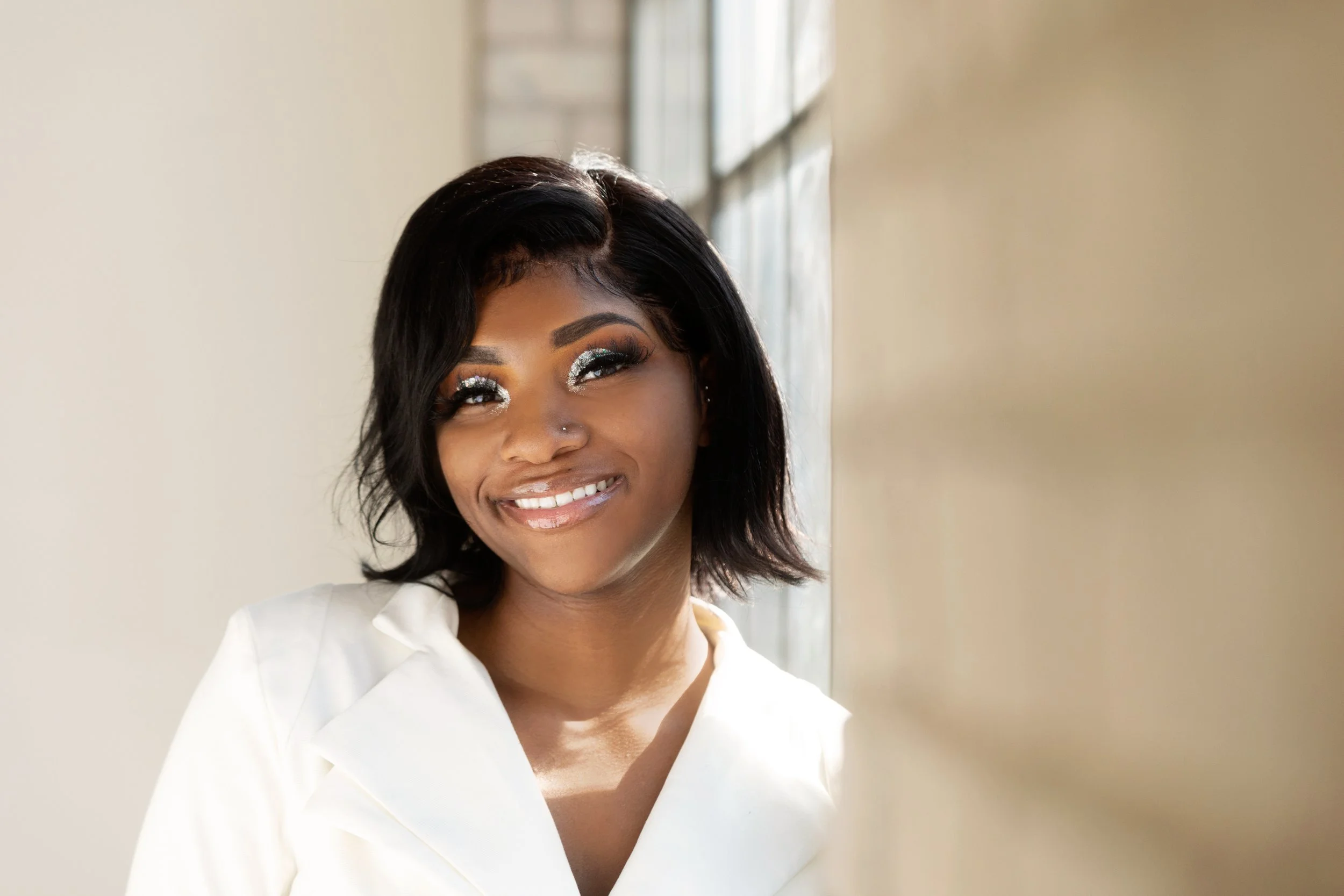A smiling woman with dark hair, silver eye makeup, and a white outfit standing near a window with sunlight.