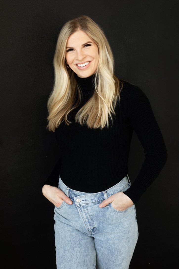A young woman with long blonde hair smiling, wearing a black turtleneck sweater and light blue jeans against a dark background.