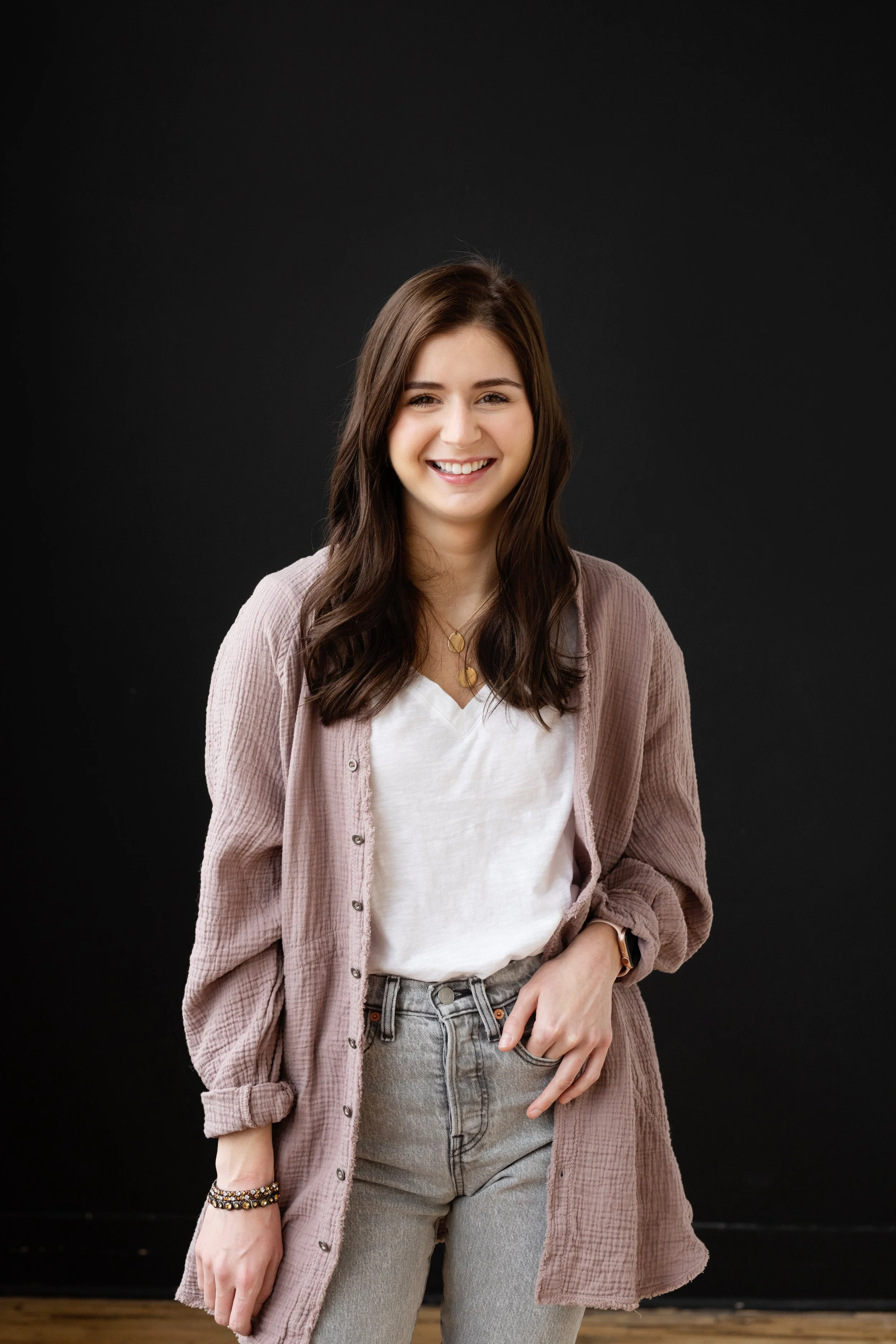 Young woman with brown hair smiling, wearing a light pink cardigan, white t-shirt, and gray jeans, standing against a black background.