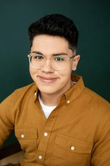 Young man with dark hair, glasses, and a friendly smile, wearing a mustard-colored button-up shirt against a dark green background.