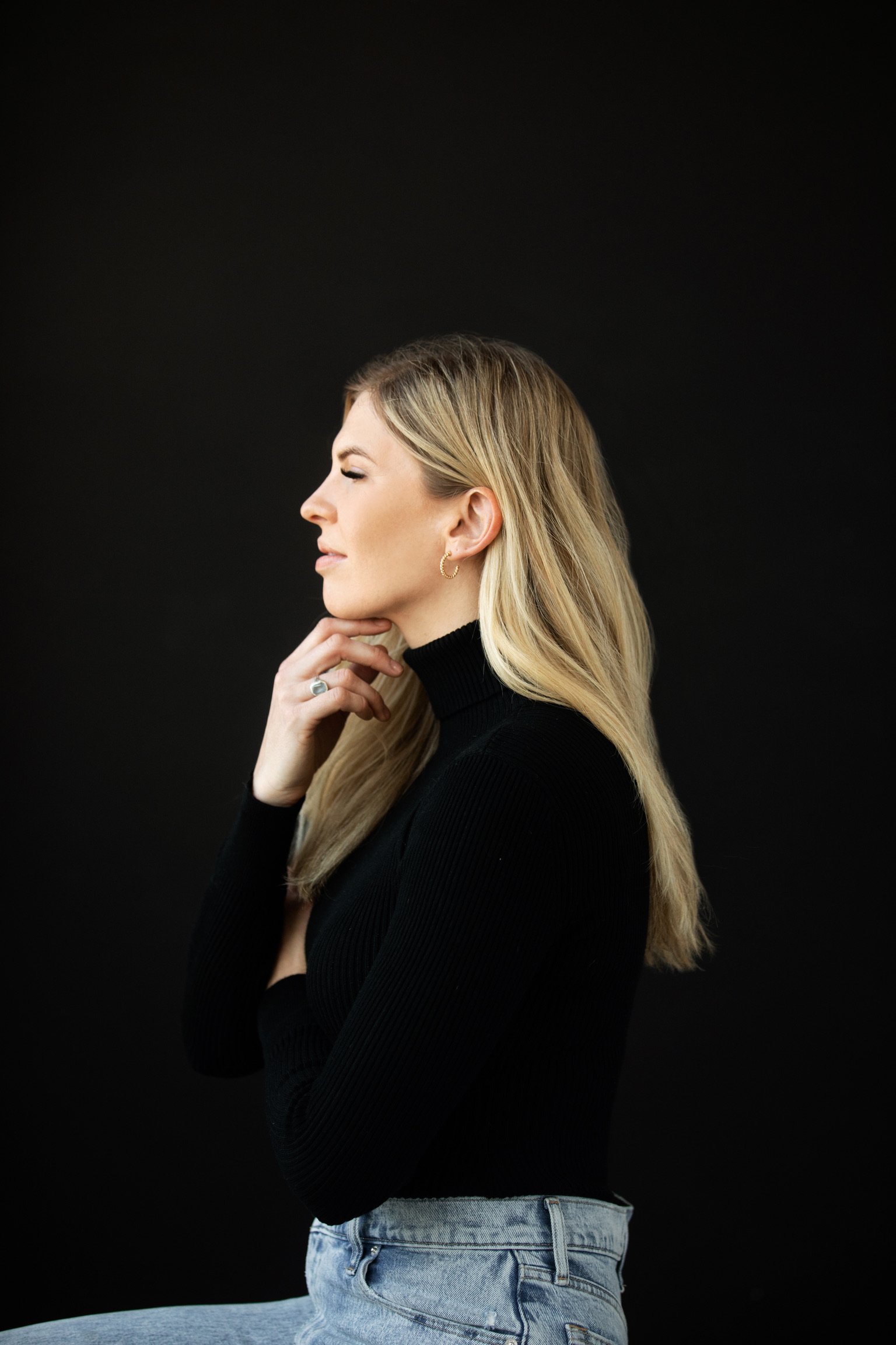 A woman with blonde hair wearing a black turtleneck and blue jeans, posing in profile against a black background.