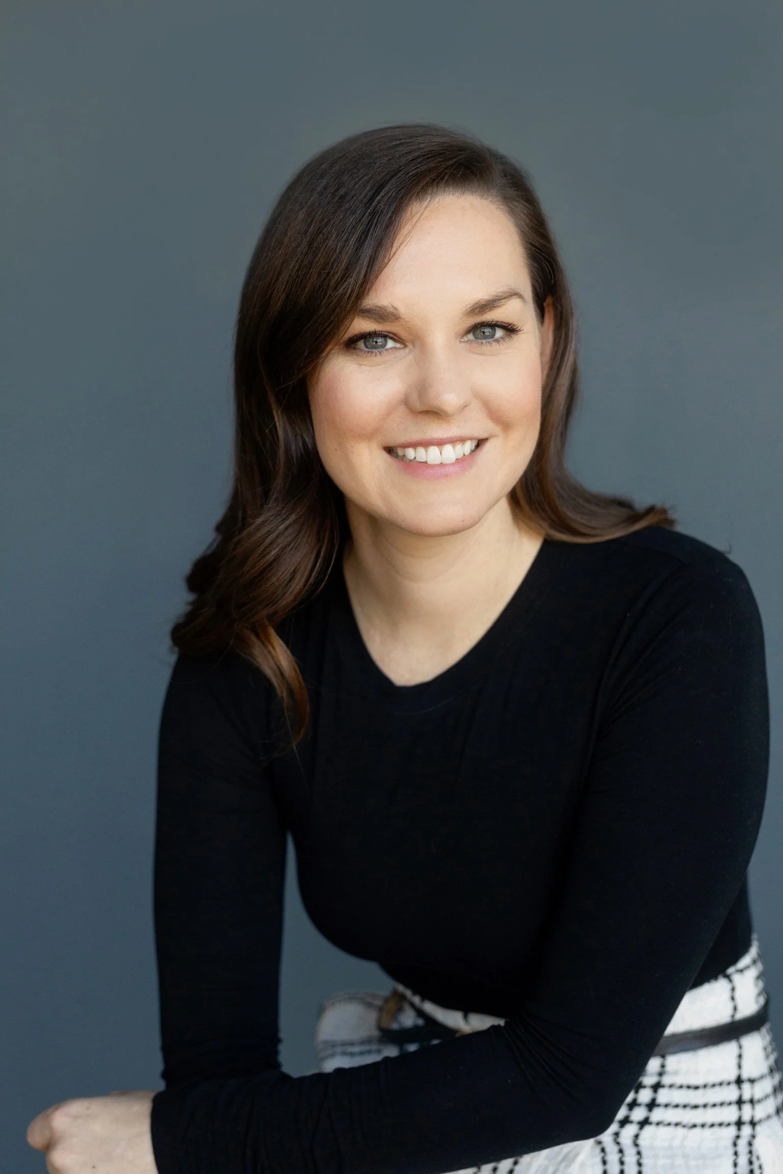 Portrait of a smiling woman with shoulder-length brown hair, wearing a black top and patterned skirt against a blue background.