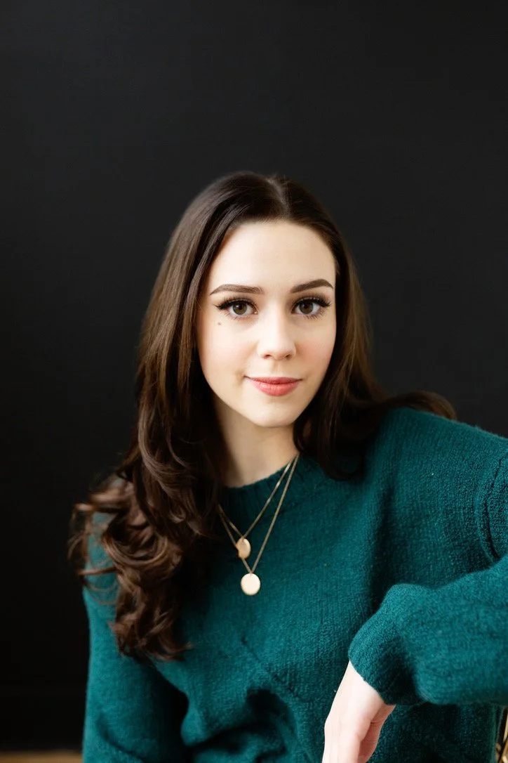 A young woman with brown hair and green eyes taking a selfie against a black background. She is wearing a teal sweater and layered necklaces.