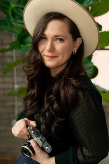 A woman with long wavy brown hair, wearing a wide-brimmed hat and a dark top, holding a camera, standing in front of green plants.