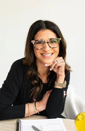 A woman with long dark hair, wearing glasses with a tortoise shell pattern, smiling and resting her chin on her hand. She is dressed in a black top and accessories, seated at a desk with a notebook and a pen, with a yellow object partially visible in