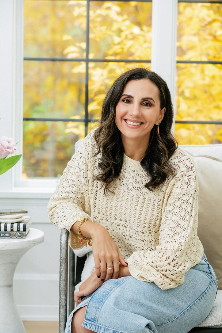 A woman with dark, wavy hair sitting in a cozy room with a large window showing autumn leaves outside. She is smiling, wearing a cream crochet sweater, accessorized with bracelets, and sitting on a white couch.