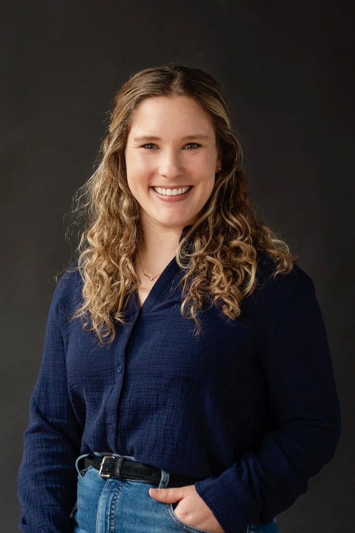 Young woman with curly blonde hair smiling and wearing a navy blue shirt and jeans against a dark background.