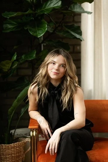 A young woman with blonde, wavy hair sitting on an orange chair indoors, with a large green plant and a window with white curtains in the background.