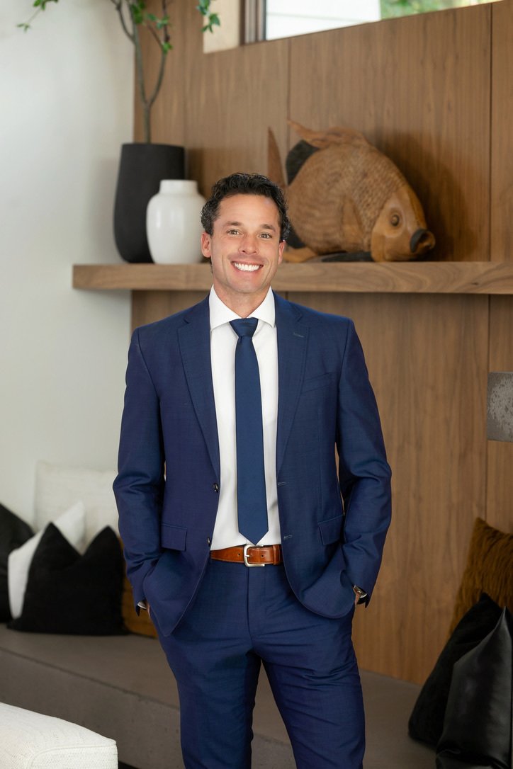A smiling man in a blue suit and tie standing in a modern room with wood wall decor and decorative items, including a large fish sculpture.