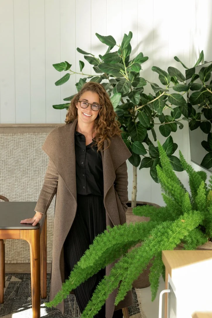 A woman with curly hair and glasses, smiling, standing near a table and large green plants inside a well-lit room.