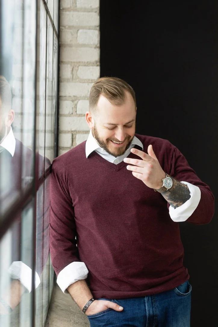 A man with a beard and tattoo on his left forearm is smiling while looking down, wearing a white collared shirt under a maroon sweater with rolled-up sleeves, standing next to a window with a brick wall behind him.