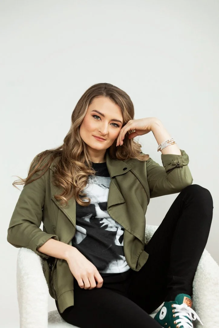 Young woman sitting on a white chair against a plain background, wearing a green jacket, graphic t-shirt, black pants, and teal sneakers, with a confident expression and relaxed pose.