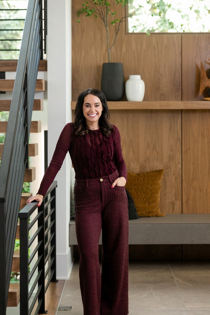 A woman with dark hair and a cheerful smile standing near a staircase inside a modern home. She is wearing a matching burgundy top and wide-leg trousers, with one hand in her pocket and the other resting on the staircase railing. The background featu