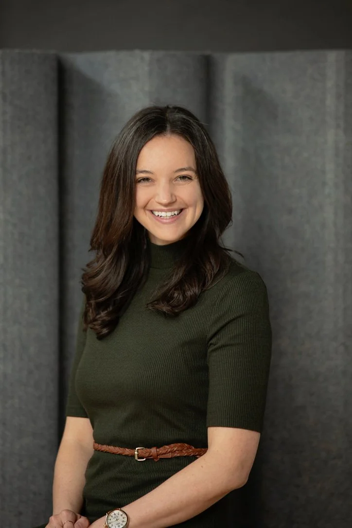 A smiling woman with long dark hair wearing a dark green short-sleeve top and a brown belt, standing in front of a grey upholstered background.