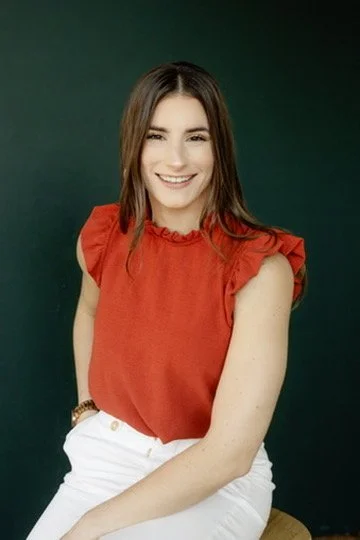 A young woman with long brown hair, smiling, wearing a red sleeveless top and white pants, sitting against a dark green background.