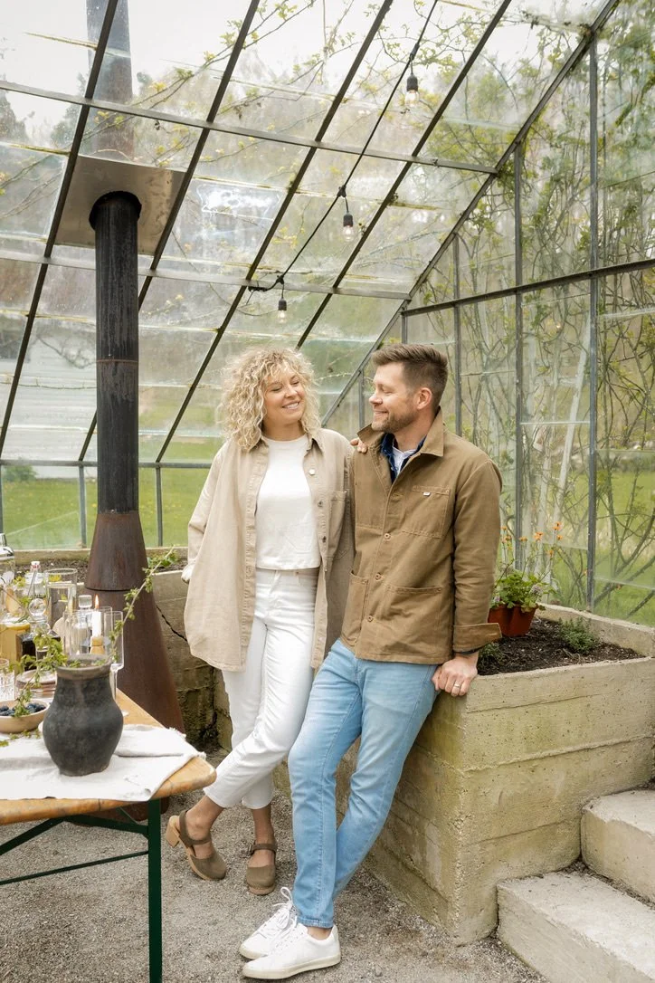 A man and woman smiling and talking inside a greenhouse with transparent walls and ceiling, with plants and gardening supplies nearby.