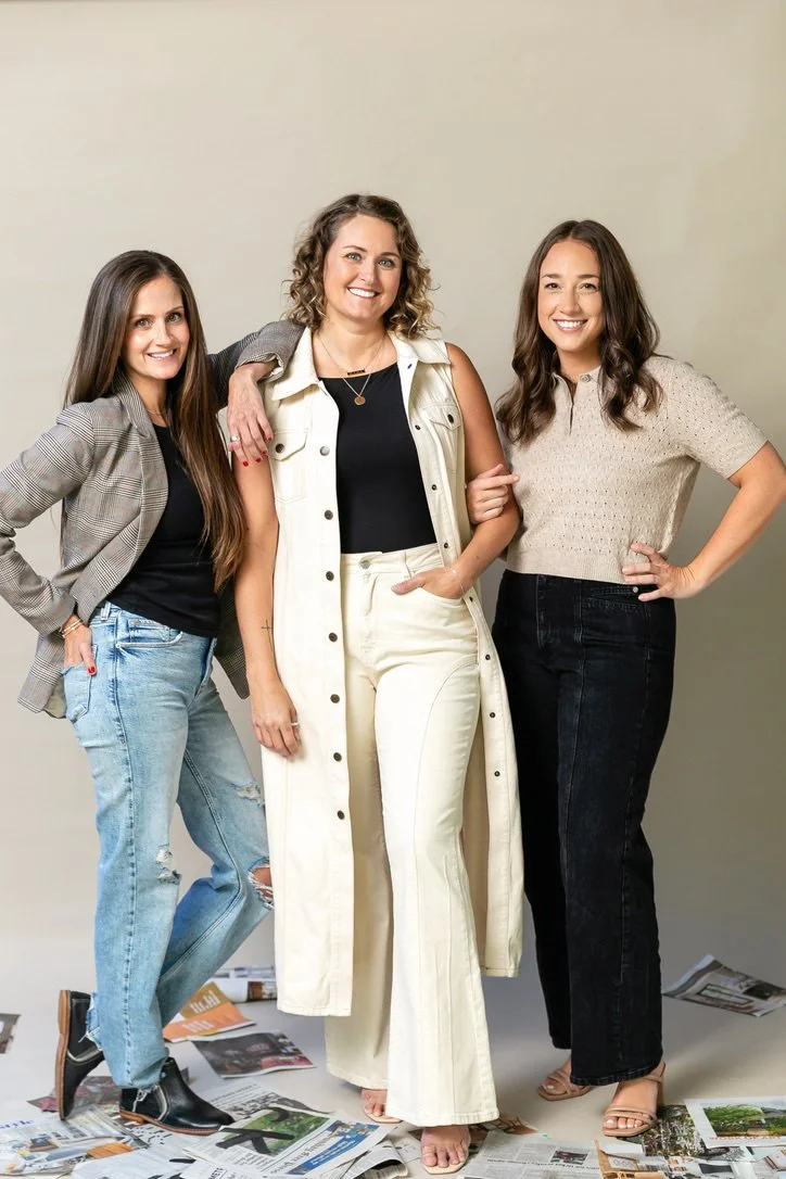 Three women standing together, smiling, with scattered magazines on the floor.